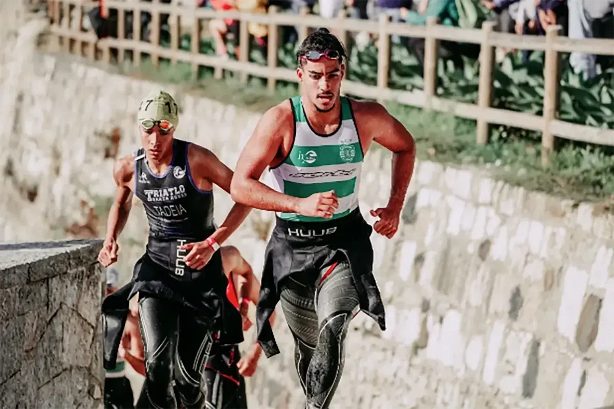 Zwei Athleten laufen bergauf, die Triathlon-Reihenfolge im Blick. Der vordere trägt ein grünes und weißes Shirt, der hintere ein schwarzes mit der Aufschrift "TRIATLO".