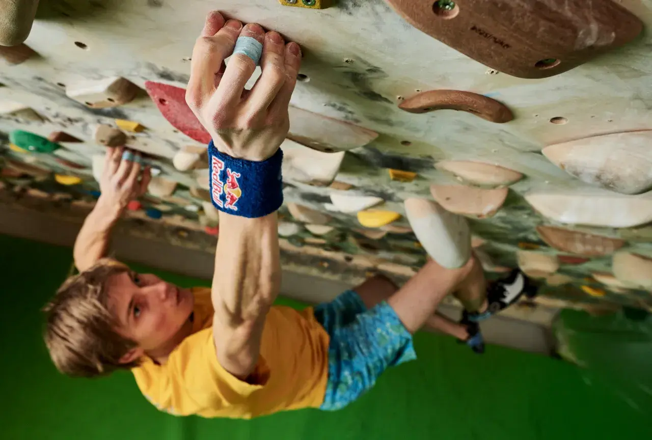 Alex Megos klettert konzentriert an einer Boulderwand. Seine Hand greift fest einen Griff, während sein Blick nach oben gerichtet ist.