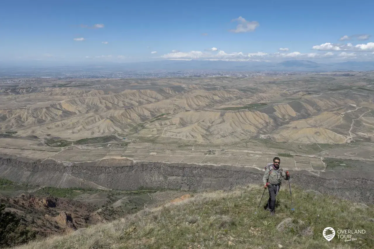 Ein Mann mit Rucksack und Wanderstöcken genießt die weite Aussicht bei seinem Armenien Wandern. Die Landschaft ist geprägt von trockenen Hügeln und einer fernen Stadt.