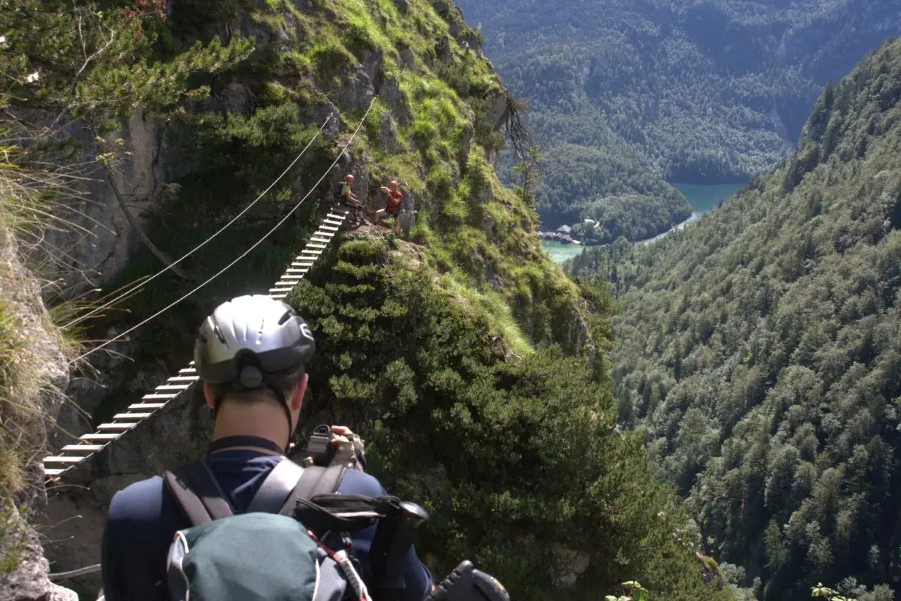 Ein Mann mit Helm und Rucksack fotografiert einen Klettersteig in Berchtesgaden. Eine Hängebrücke führt über eine Schlucht mit Blick auf einen See.
