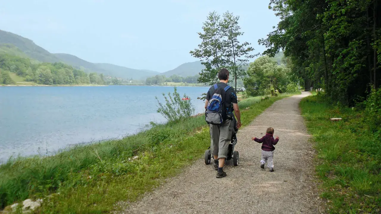 Vater schiebt Kinderwagen am Happurger Stausee entlang, während das Kind vorausläuft. Herrliche Kulisse für eine Wanderung.