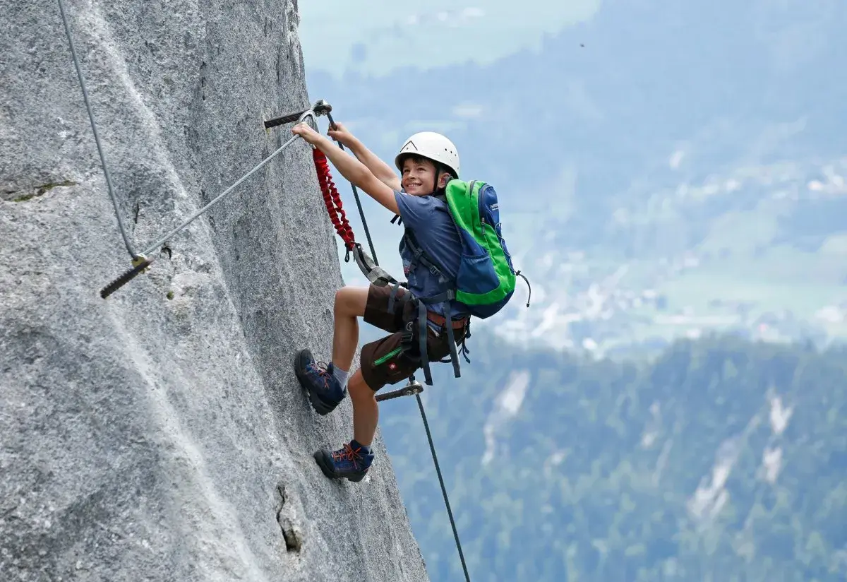 Ein Junge mit Helm und Rucksack klettert einen Felsgrat. Ein spannendes Abenteuer für klettersteig kinder.