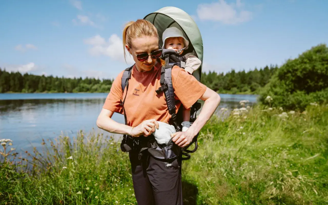 Frau mit kraxe baby am Seeufer, umgeben von grüner Natur und Bäumen.