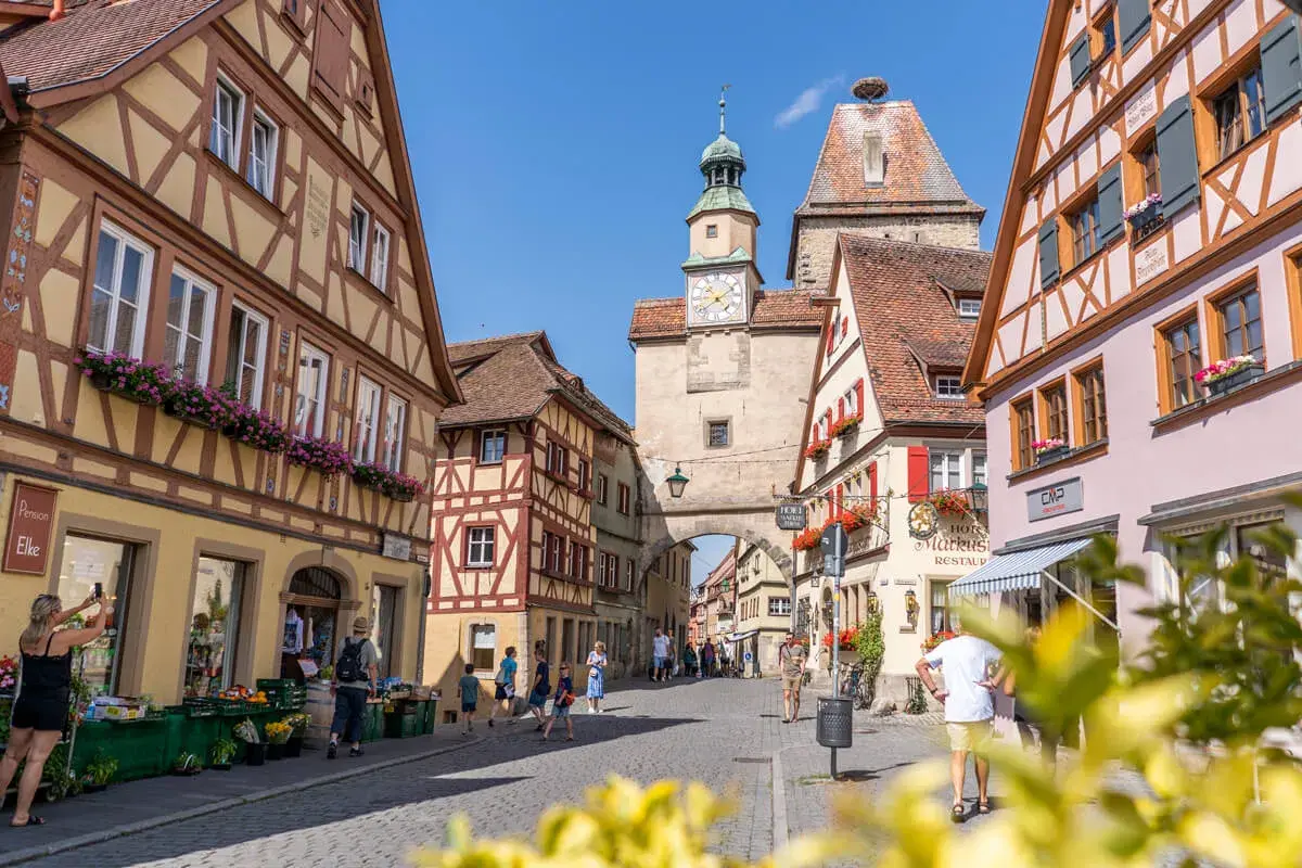 Fachwerkhäuser säumen die Gasse in Rothenburg ob der Tauber. Ein Uhrenturm überragt die Szene, während Touristen die mittelalterliche Stadt erkunden.