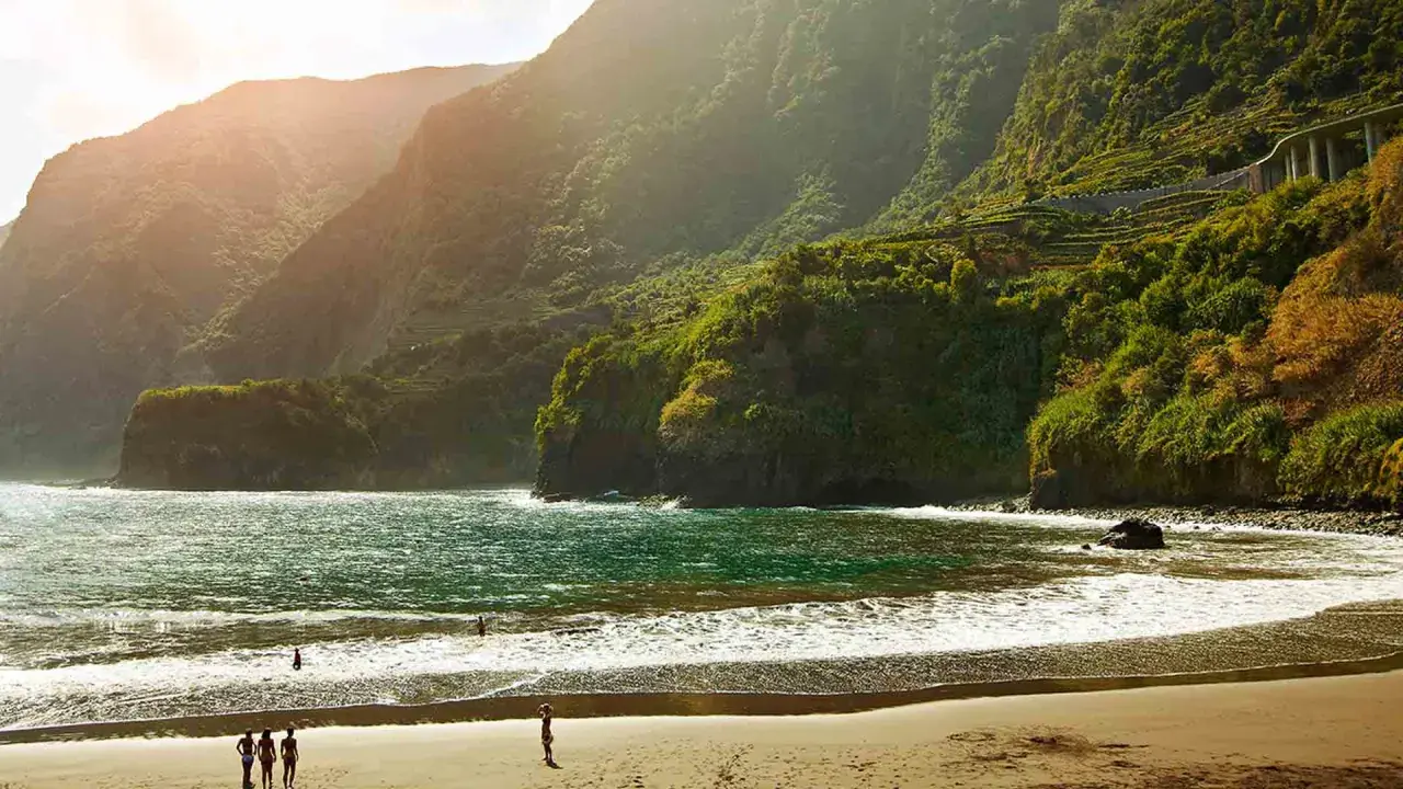 Sonne beleuchtet den malerischen **Madeira schwarzer Strand** mit üppig grünen Bergen im Hintergrund. Wellen brechen sanft am Ufer.