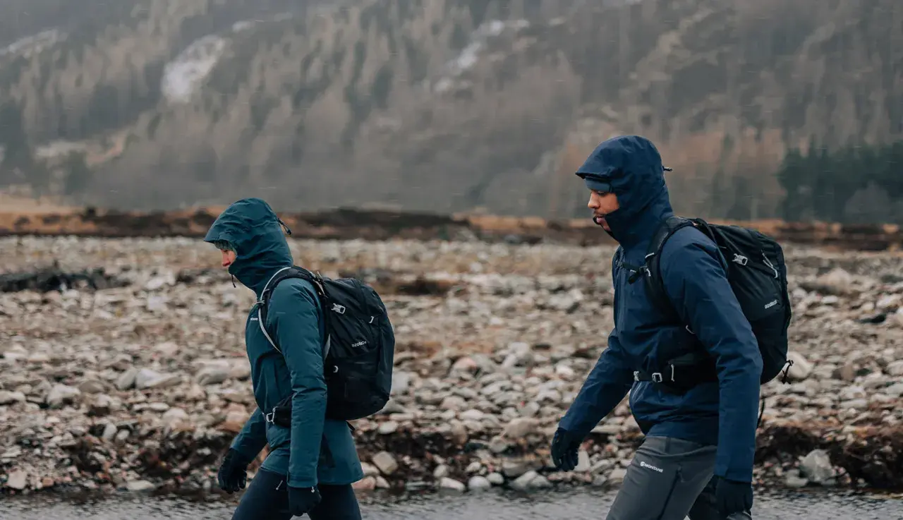 Zwei Wanderer mit Rucksäcken gehen durch eine steinige Landschaft in Schottland. Das Wetter ist regnerisch, die Kapuzen sind hochgezogen.