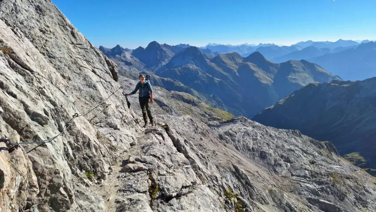 Frau auf dem **Heilbronner Weg wandern**, gesichert an einem Stahlseil, mit atemberaubendem Bergpanorama im Hintergrund.