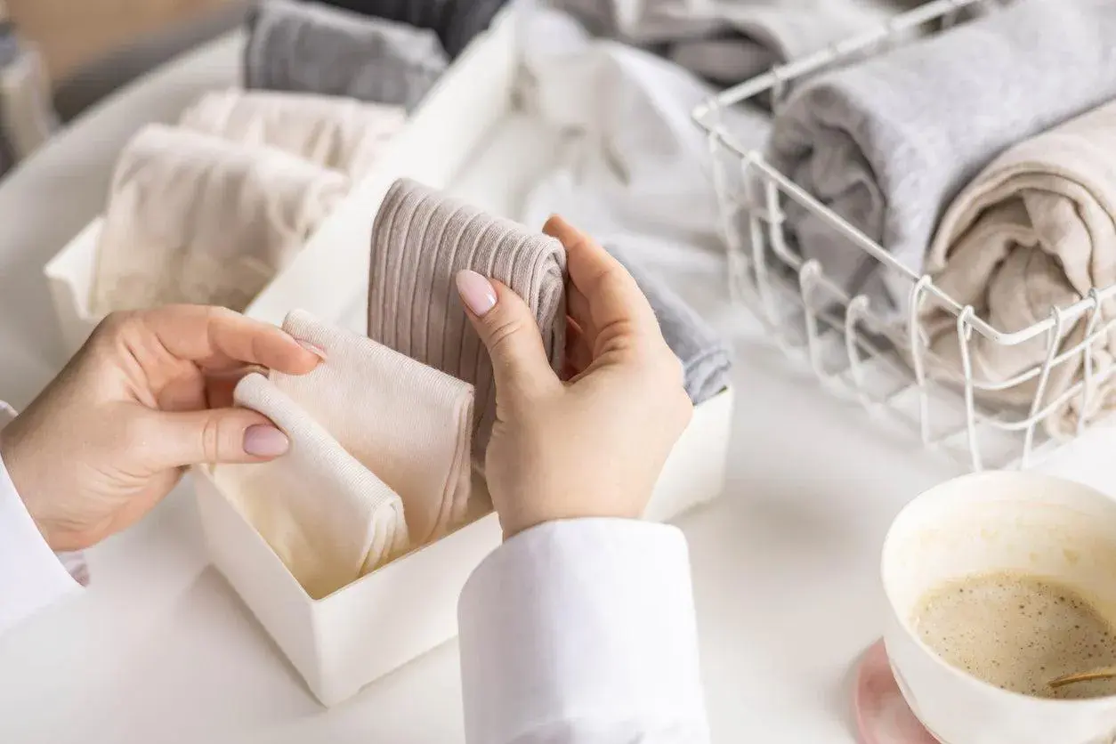 Hände sortieren gefaltete Kleidung, darunter auch warme **Wandersocken**, in einem Behälter. Daneben ein Korb mit weiteren Textilien und eine Kaffeetasse.