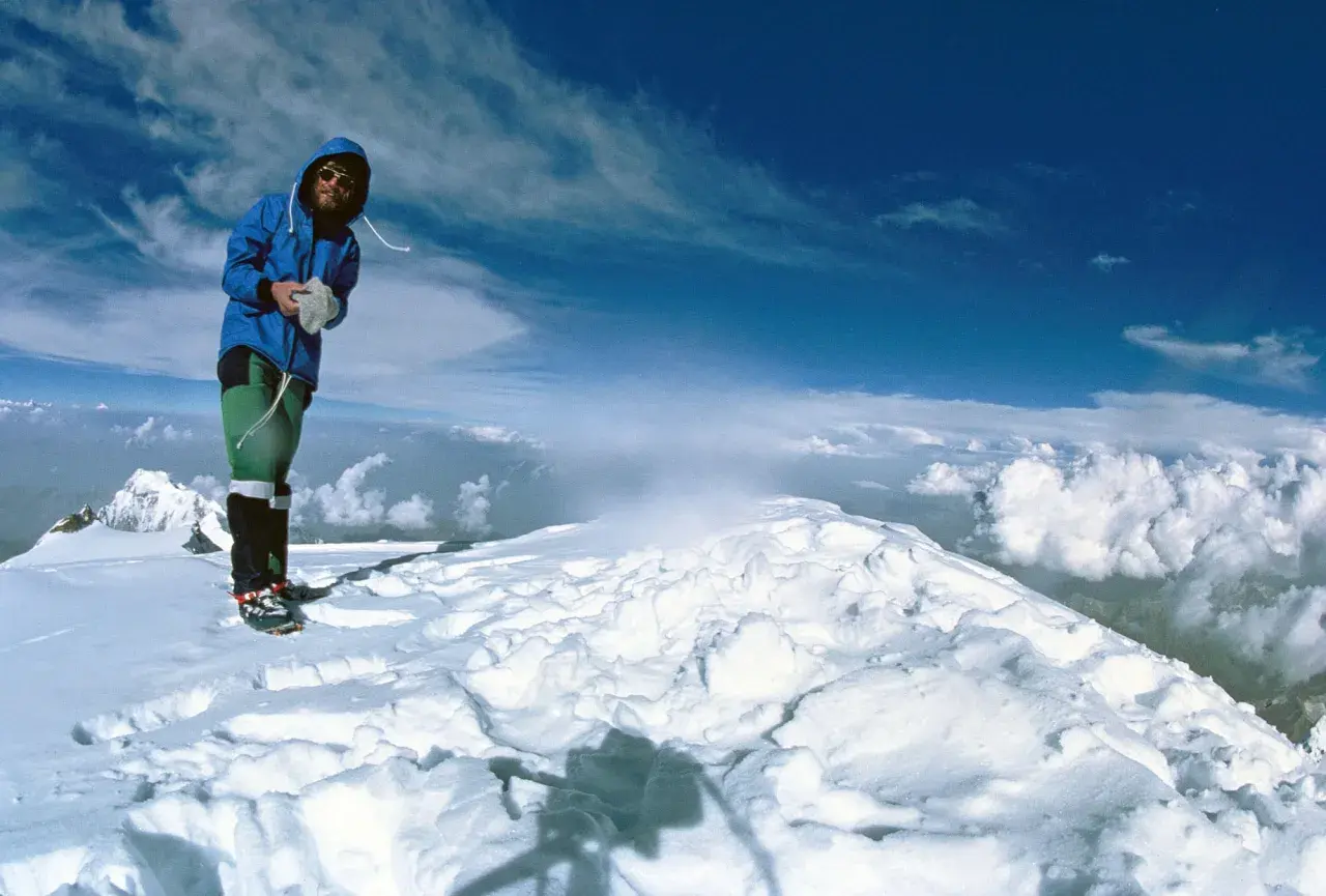 Reinhard Messner steht auf einem schneebedeckten Gipfel, umgeben von Wolken und blauem Himmel.