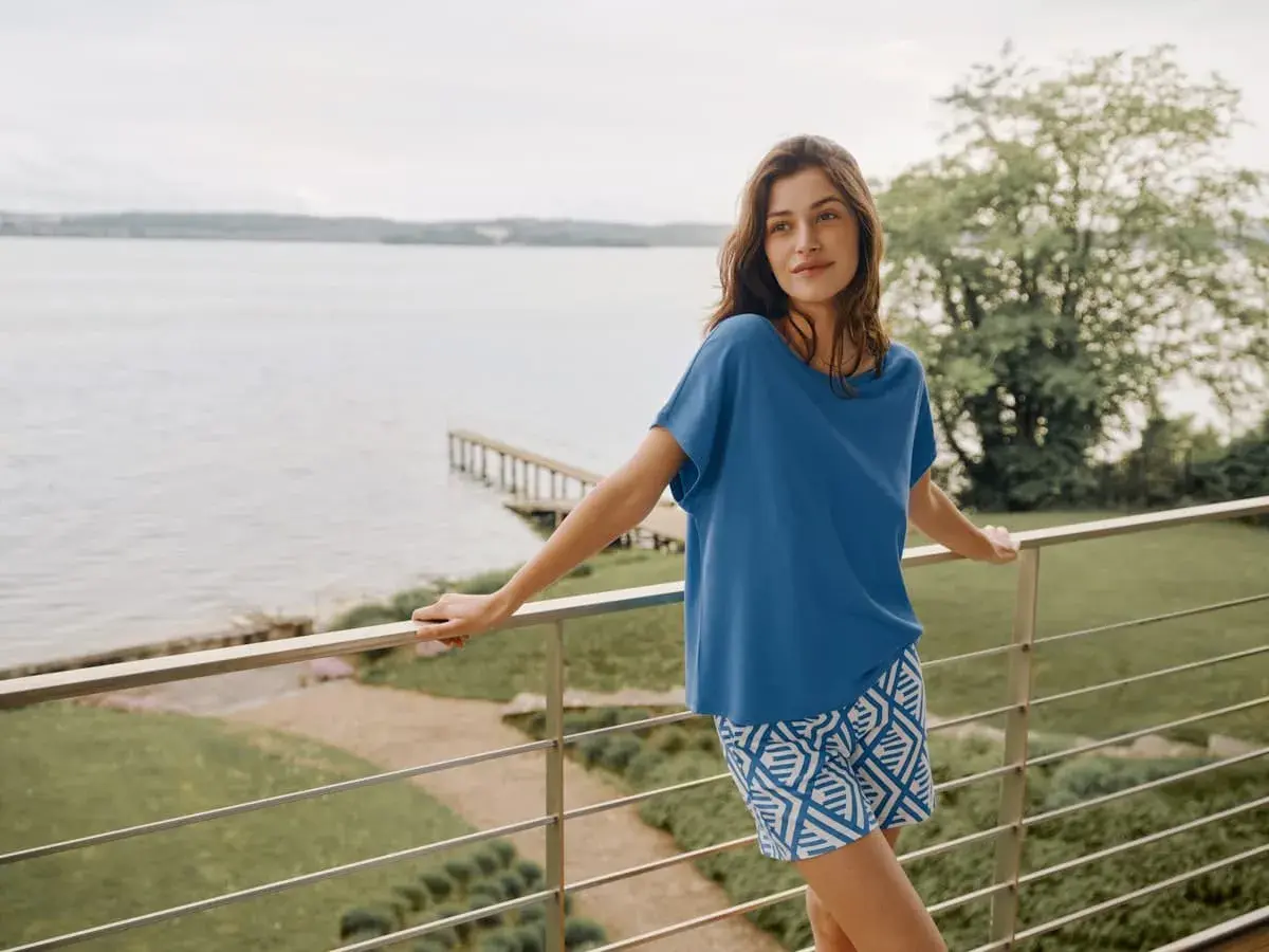 Frau auf Balkon mit Blick auf See, trägt blaues Oberteil aus modal stoff und gemusterte Shorts.