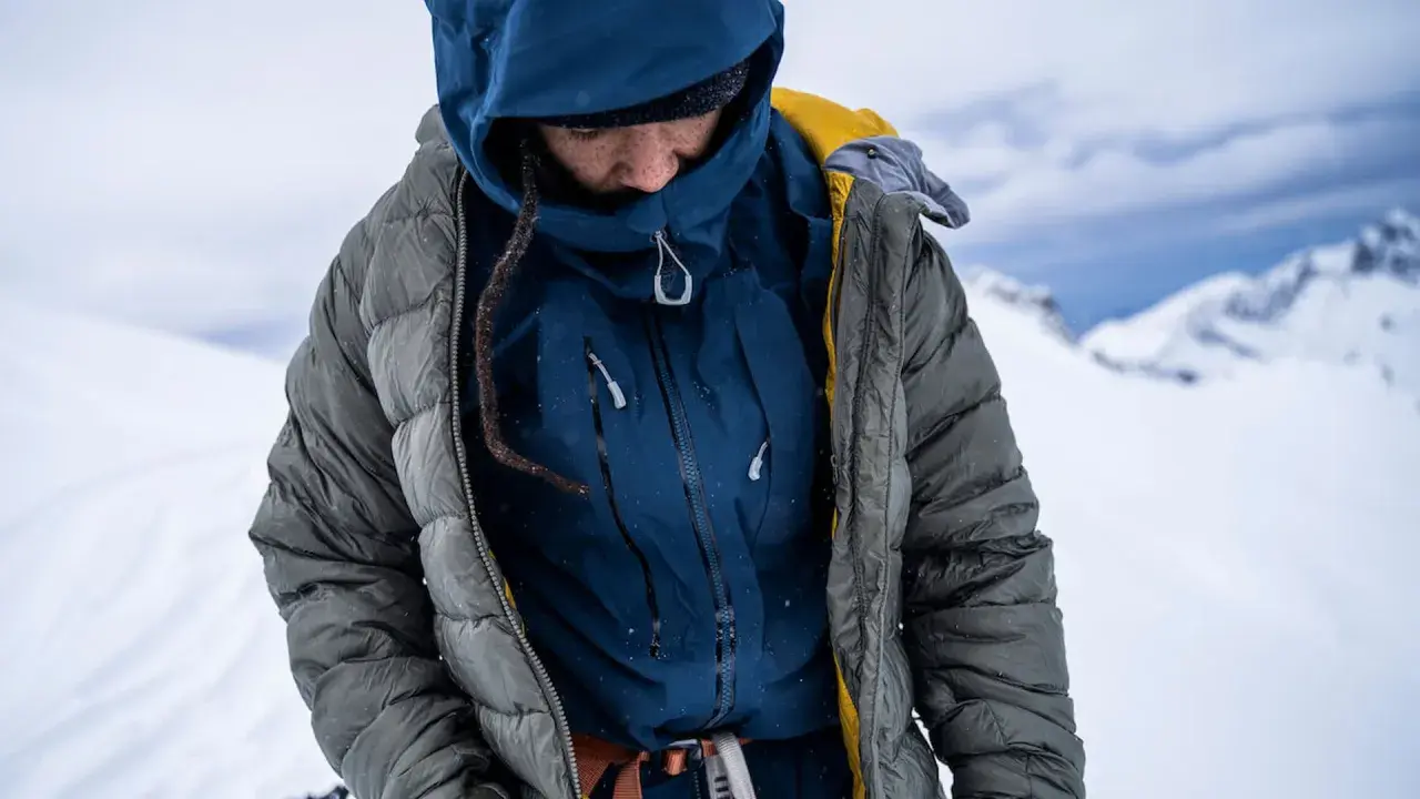 Person in Daunenjacke im Schnee. Die Jacke ist grau, darunter eine blaue Schicht. Perfekt für das Daunenjacke waschen nach einer Bergtour.