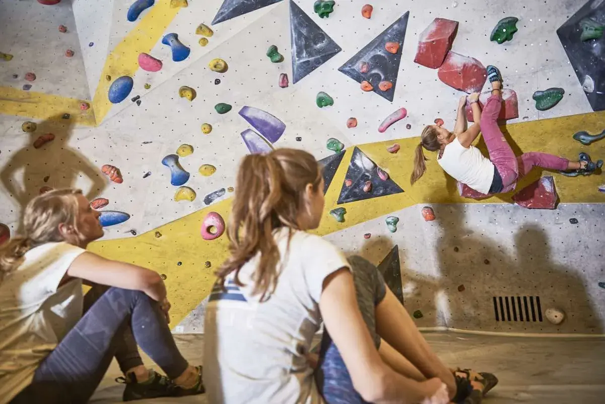 Zwei Mädchen schauen zu, wie eine dritte Person an der Kletterwand hochklettert. Sie tragen bequeme Kleidung, wie man sie zum Bouldern anzieht: Leggings und T-Shirts.