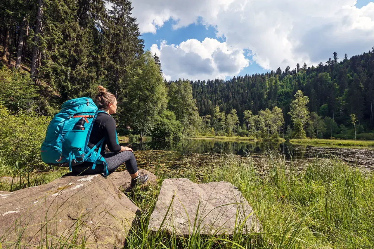 Frau mit Rucksack sitzt an einem See, umgeben von Wald. Ein perfekter Moment auf einer der vielen **weitwanderwege europa**.