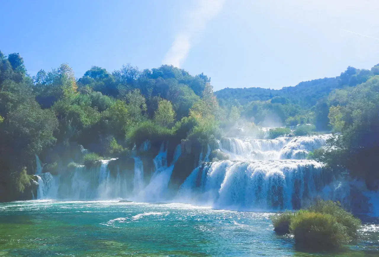 Ein majestätischer Krk Wasserfall stürzt über mehrere Stufen in ein türkisfarbenes Becken. Üppige grüne Bäume säumen die Klippen und der Himmel ist strahlend blau.