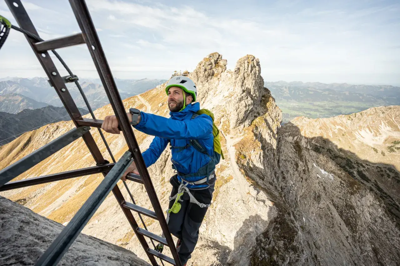 Mann klettert auf einem Leiternabschnitt des Oberstdorf Klettersteigs. Die Aussicht auf die Alpen ist atemberaubend.