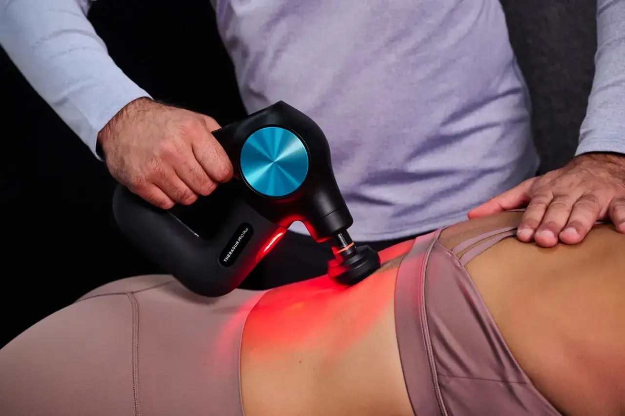 Eine Hand führt eine Massagepistole zur Anwendung am unteren Rücken einer Person. Rotes Licht leuchtet auf.