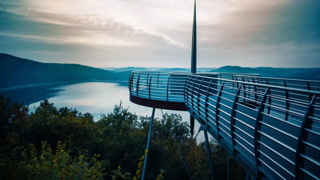 Moderne Aussichtsplattform über einem See, ideal für das Wandern in NRW. Sanfte Hügel und ein dramatischer Himmel im Hintergrund.