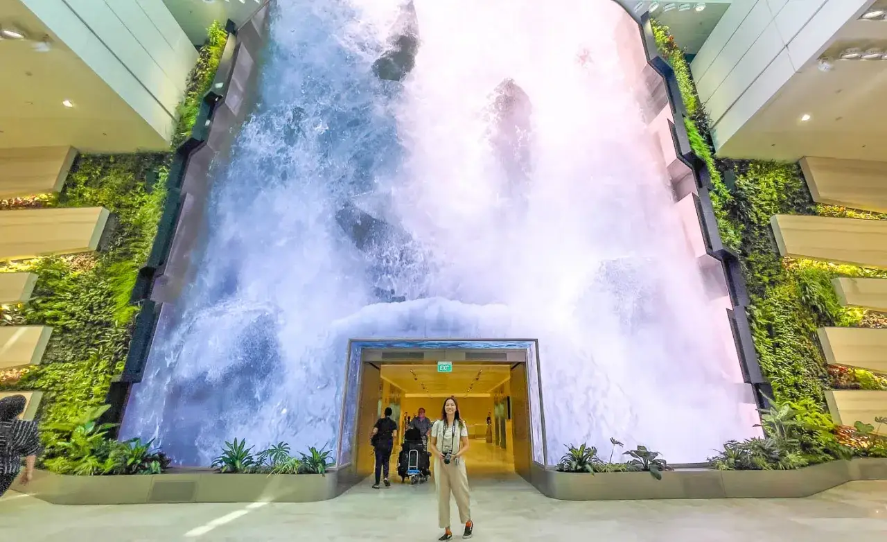 Frau steht vor einer riesigen digitalen Wasserfallwand im Singapur Flughafen. Gr&uuml;ne Pflanzen schm&uuml;cken die Seiten.