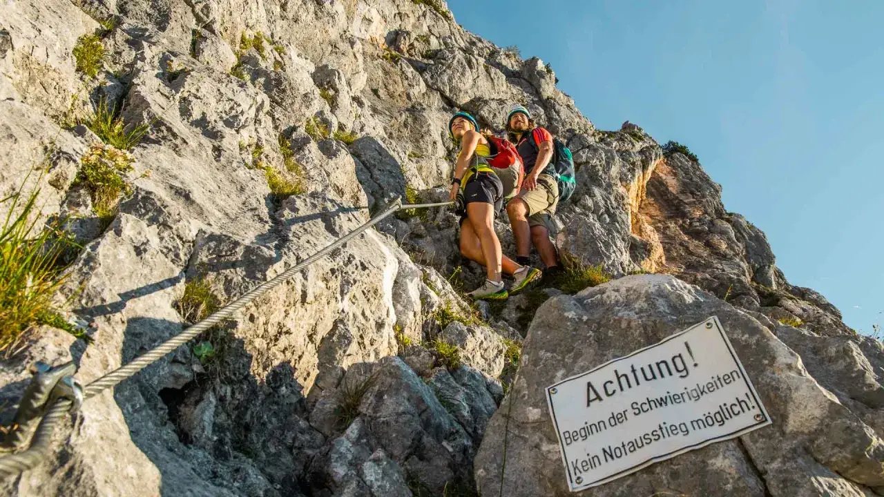Zwei Wanderer am Klettersteig Berchtesgaden, gesichert an einem Stahlseil. Ein Schild warnt vor Schwierigkeiten.