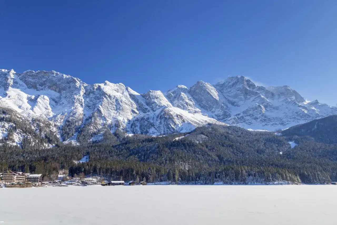 Eibsee winter: Majest&auml;tische, schneebedeckte Berge &uuml;ber einem zugefrorenen See mit H&auml;usern am Ufer.