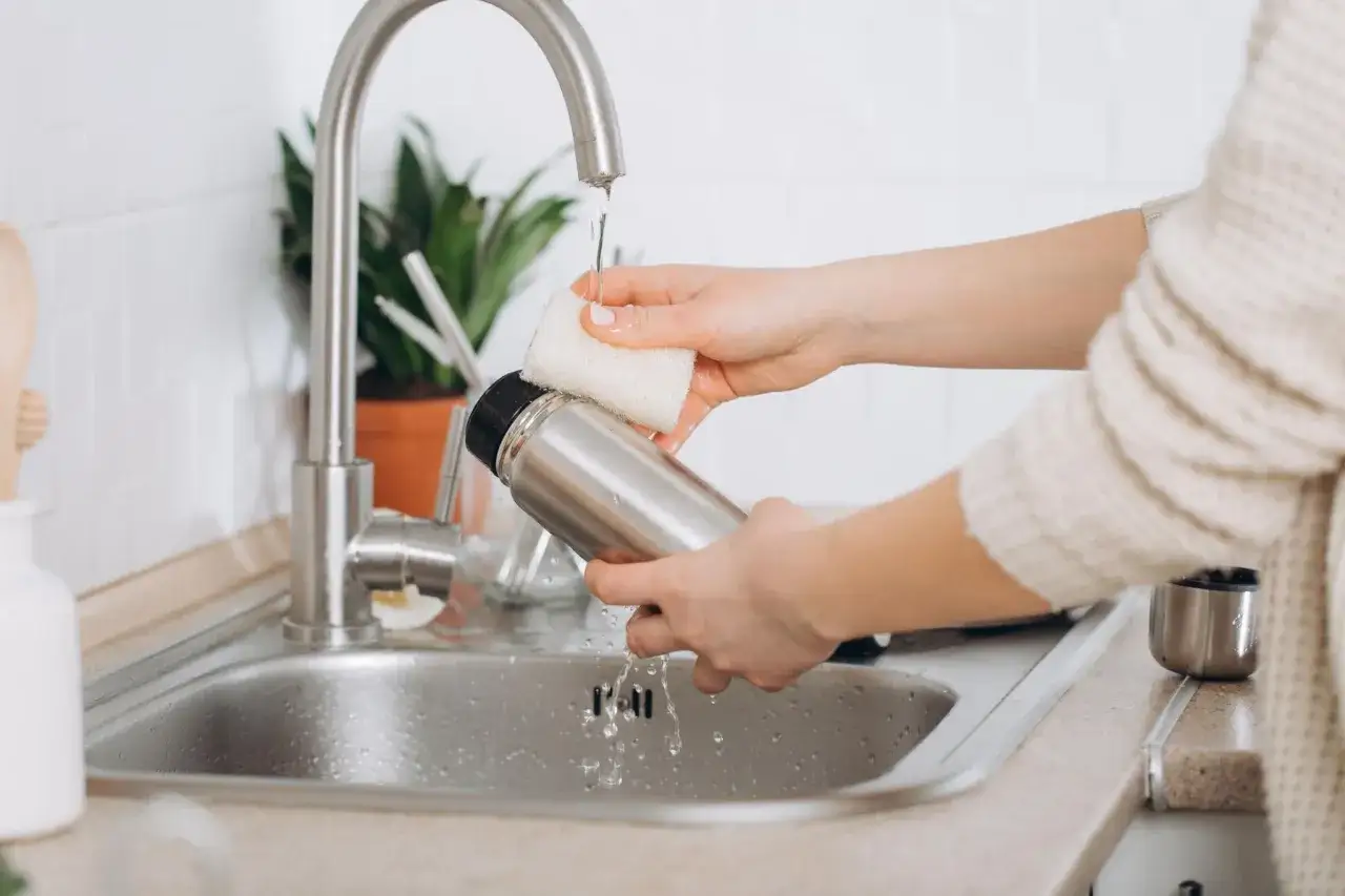 H&auml;nde reinigen eine Thermoskanne unter flie&szlig;endem Wasser in der Sp&uuml;le.