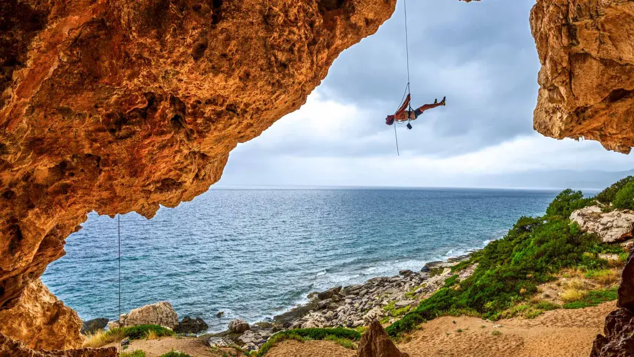 Ein Kletterer schwebt an einem Seil vor der K&uuml;ste Sardiniens. Das Meer glitzert unter einer dramatischen Felsformation.