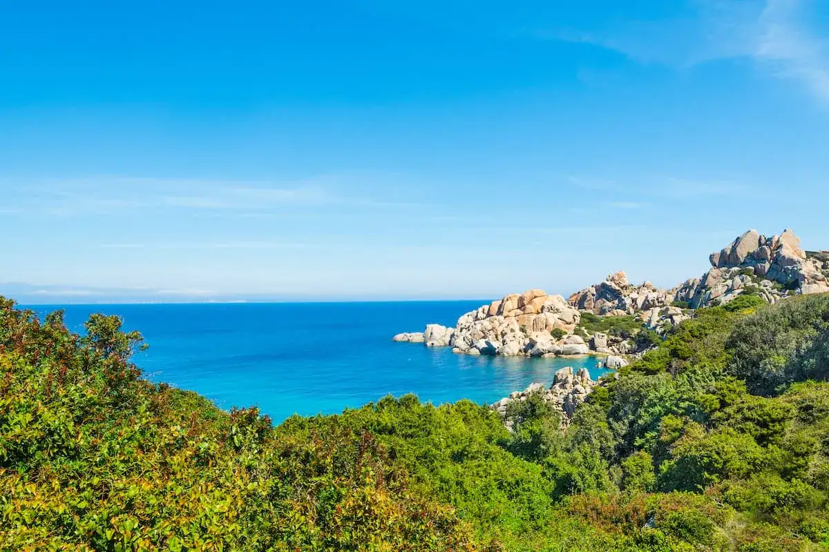 Blick auf das Meer und die Felsen von Capo Testa. Gr&uuml;ne Vegetation im Vordergrund, t&uuml;rkisfarbenes Wasser und ein klarer blauer Himmel.
