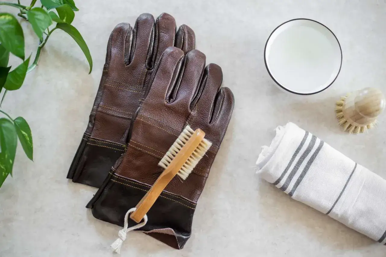Braune lederhandschuhe liegen bereit zum lederhandschuhe waschen. Eine B&uuml;rste, eine Sch&uuml;ssel mit Wasser und ein Handtuch sind daneben platziert.