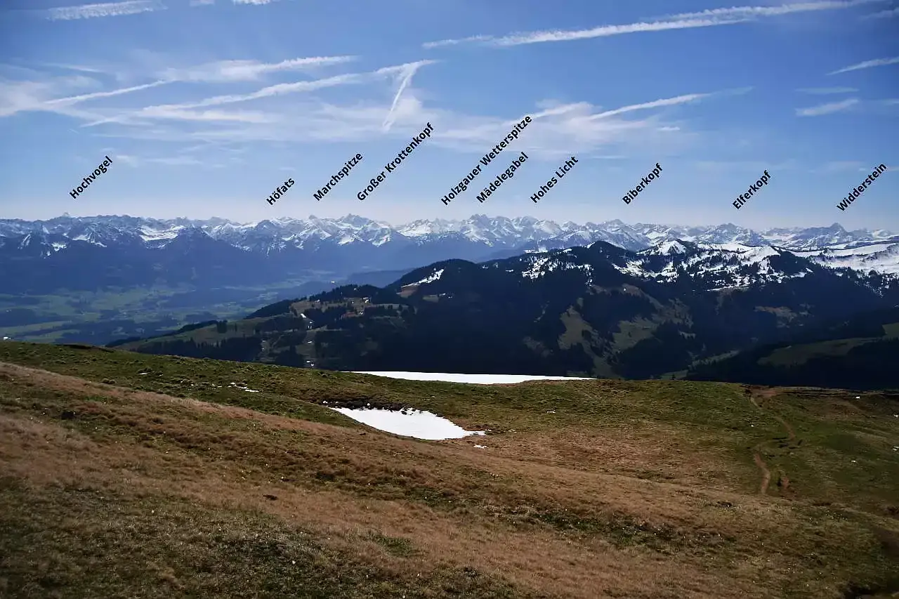 Blick über grüne Hügel und schneebedeckte Gipfel. Ein Pfad schlängelt sich den Hang hinauf, wie eine Leiter zum Steineberg.