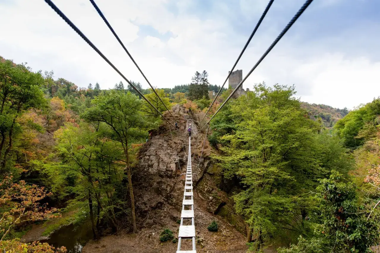 Abenteuer auf dem Klettersteig Eifel: Eine H&auml;ngebr&uuml;cke f&uuml;hrt &uuml;ber eine felsige Schlucht mit Burgruine im Hintergrund.
