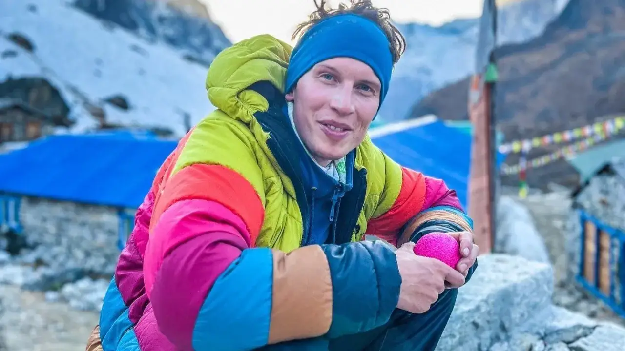 Jost Kobusch, mit einem bunten Daunenmantel und blauer M&uuml;tze, h&auml;lt einen pinken Ball in der Hand. Im Hintergrund sind schneebedeckte Berge und bunte Gebetsfahnen zu sehen.