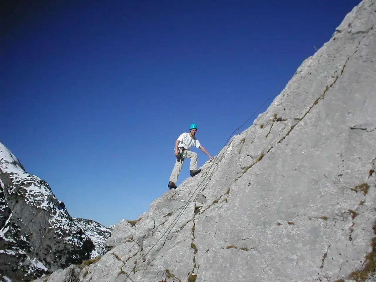 Ein Mann klettert gesichert am Seil den Tajakante Klettersteig entlang. Der blaue Himmel spannt sich über die felsige Berglandschaft.