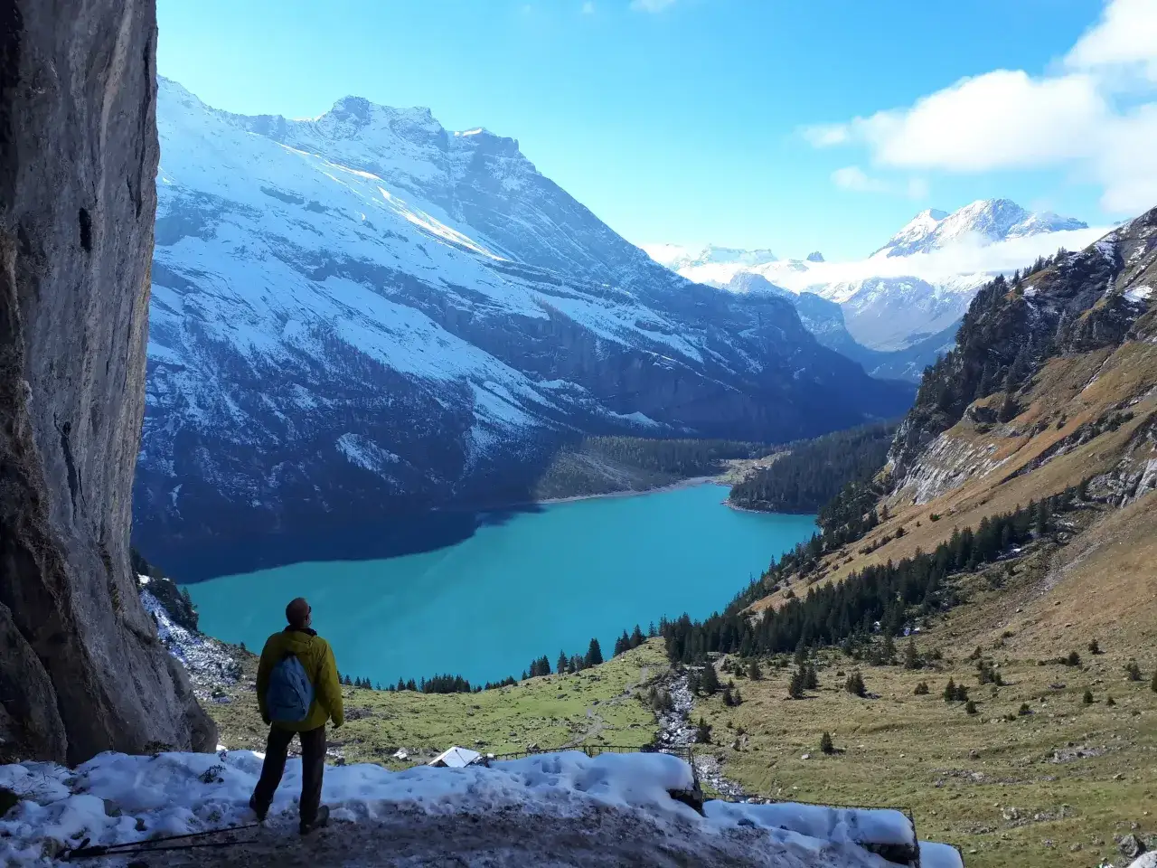 Ein Wanderer blickt auf einen t&uuml;rkisfarbenen See in den Schweizer Alpen. Schneebedeckte Berge ragen im Hintergrund auf.