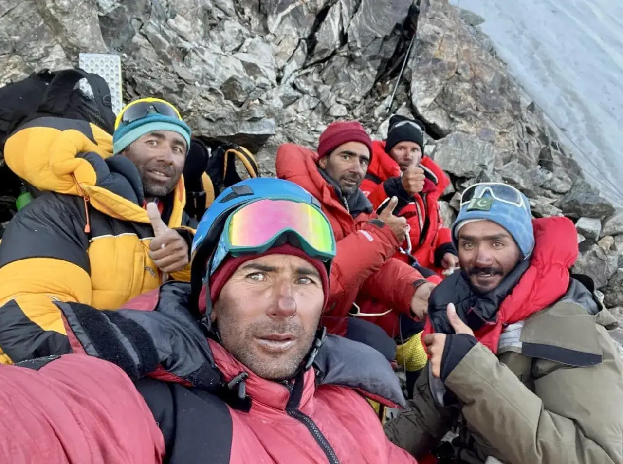 F&uuml;nf Bergsteiger, darunter Mohammed Hassan, geben Daumen hoch am K2. Sie tragen warme Kleidung und Skibrillen.