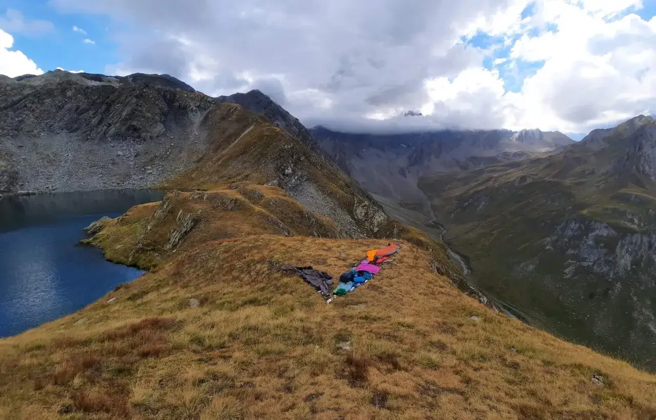 Bunte Schlafs&auml;cke liegen auf einem Bergkamm am via alpina roter weg. Ein See und steile Berge im Hintergrund.