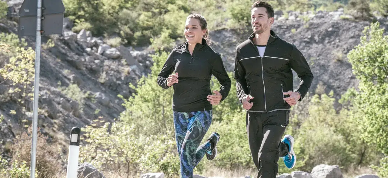 Ein Paar genie&szlig;t das Laufen im Winter, gekleidet in funktionale Sportkleidung. Sie laufen auf einem steinigen Weg, umgeben von gr&uuml;ner Vegetation.