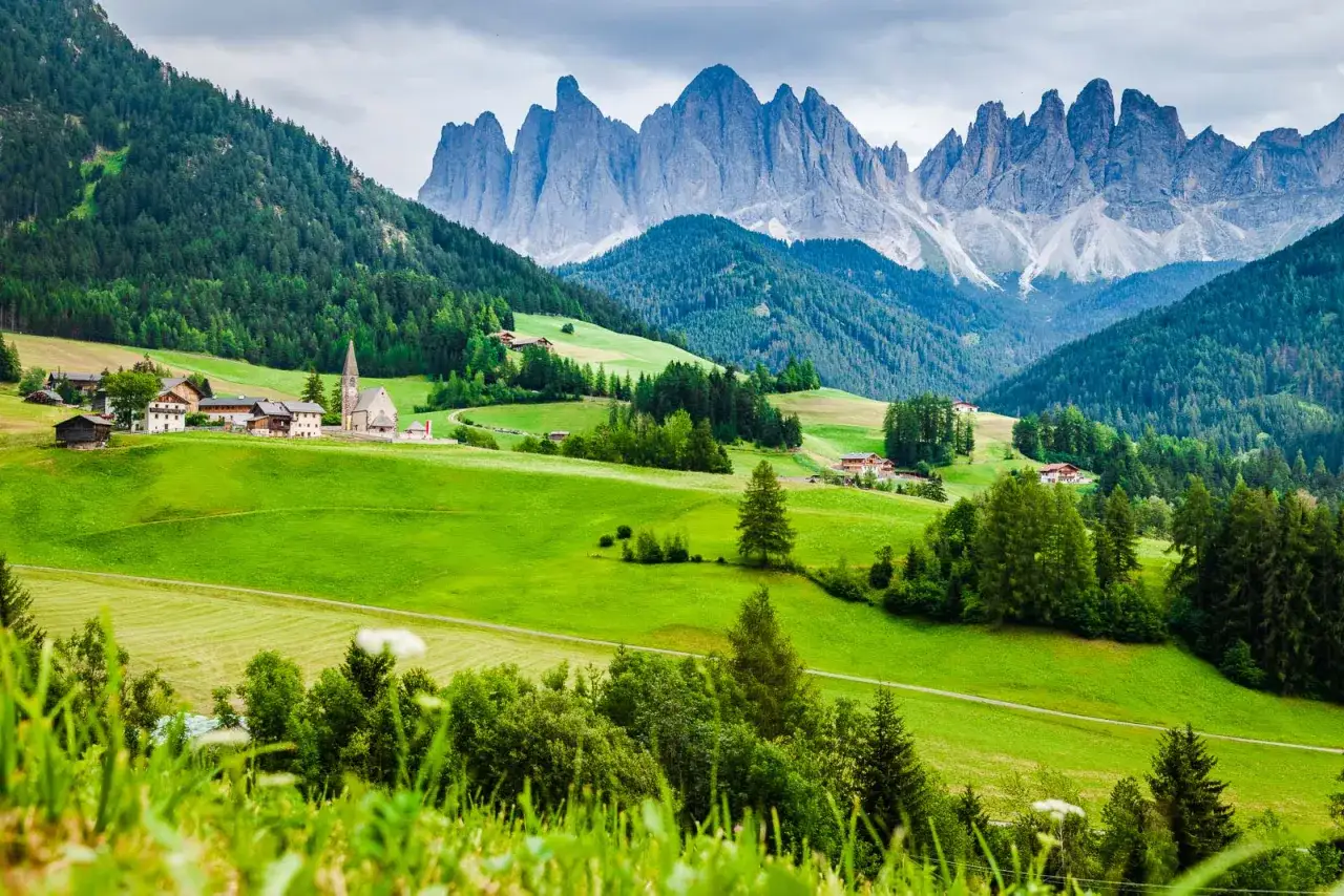 Grüne Hügel und traditionelle Häuser im Val di Funes, mit den schroffen Dolomiten im Hintergrund.