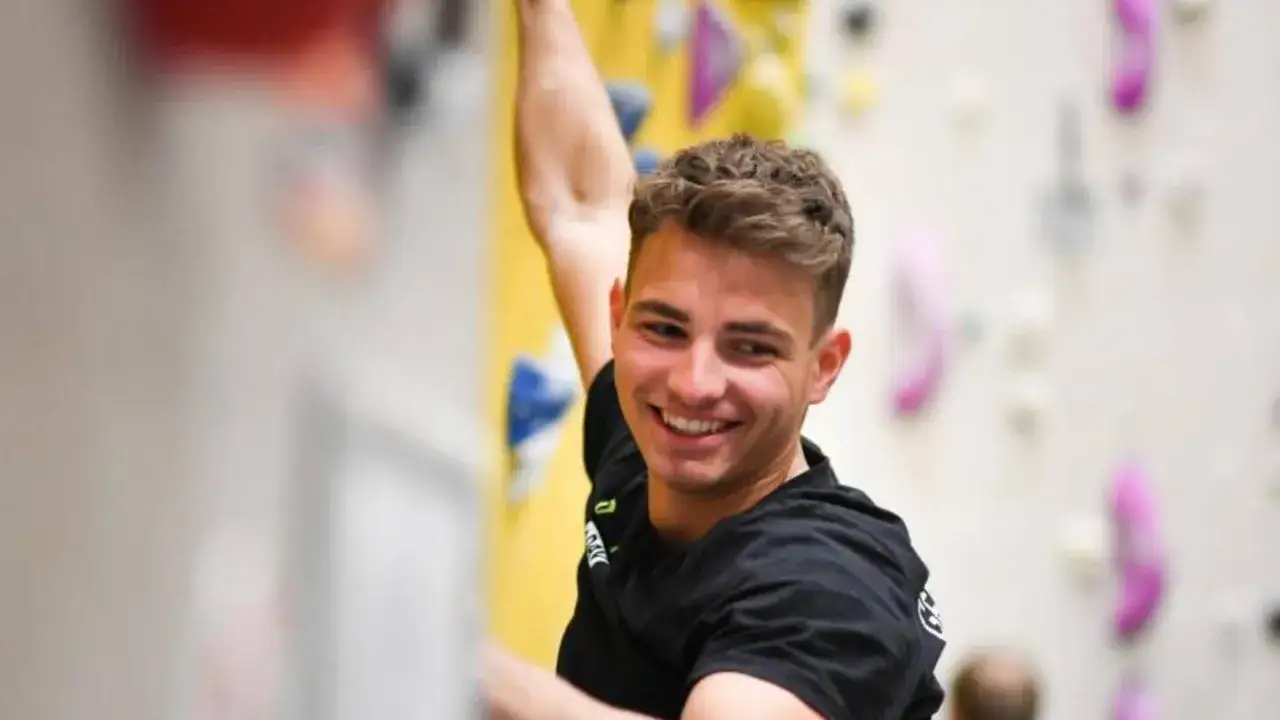 Yannick Floh&eacute; lacht, w&auml;hrend er an einer Kletterwand hochklettert. Bunte Griffe sind an der Wand befestigt.
