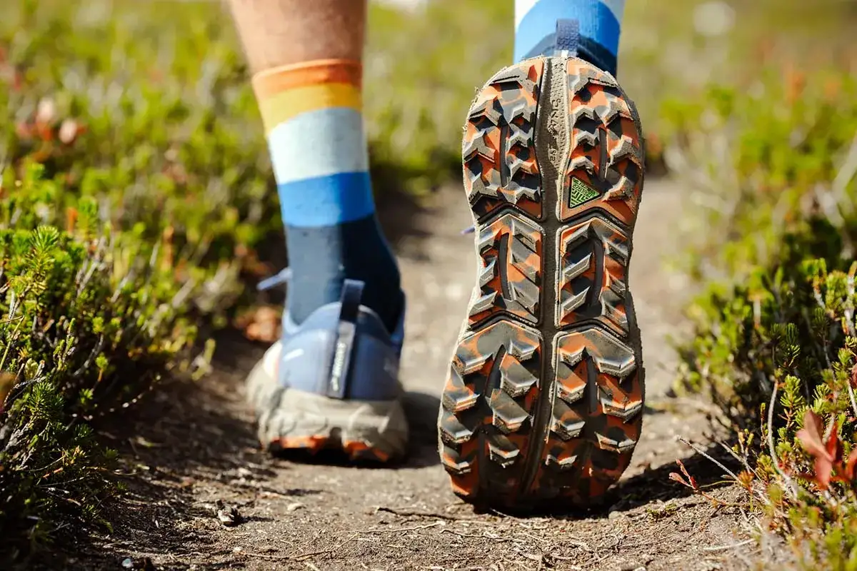 Detailaufnahme der Sohle von was sind trailrunning schuhe, die auf einem erdigen Pfad mit gr&uuml;nem Gestr&uuml;pp nebenan unterwegs sind.