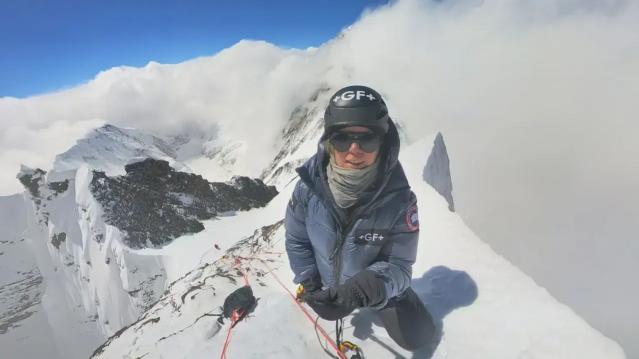 Anja Blacha auf einem schneebedeckten Berggipfel, umgeben von Wolken. Sie tr&auml;gt Bergsteigerausr&uuml;stung und blickt in die Kamera.