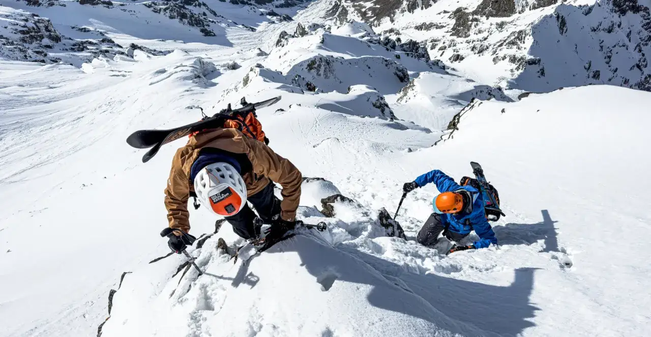 Zwei Skibergsteiger kämpfen sich durch steiles, schneebedecktes Gelände. Einer trägt ein Snowboard auf dem Rücken, der andere einen Skistock. Sie nutzen ihre Hände und Stöcke, um die rupal wand zu erklimmen.