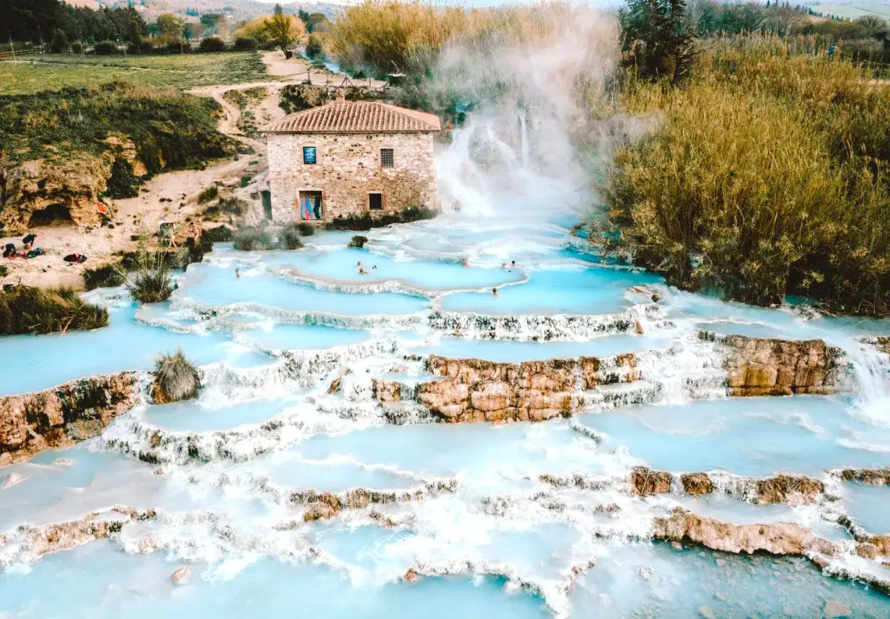 Natürliche türkisfarbene Becken und Wasserfälle bei Bagni di San Filippo, mit einem alten Steinhaus im Hintergrund.