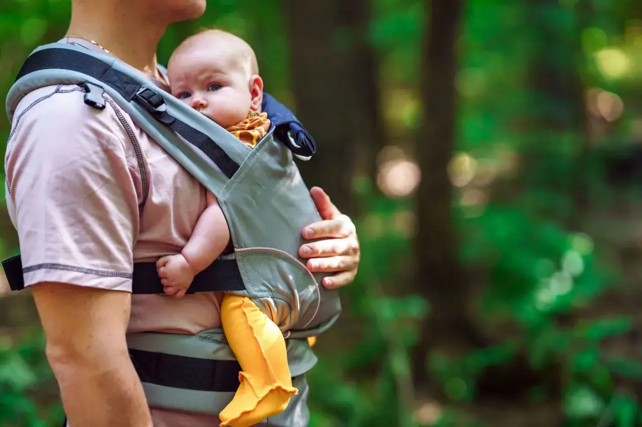 Ein Baby in einer Kraxe wird von einem Mann im Wald getragen. Das Kind schaut neugierig in die Kamera.