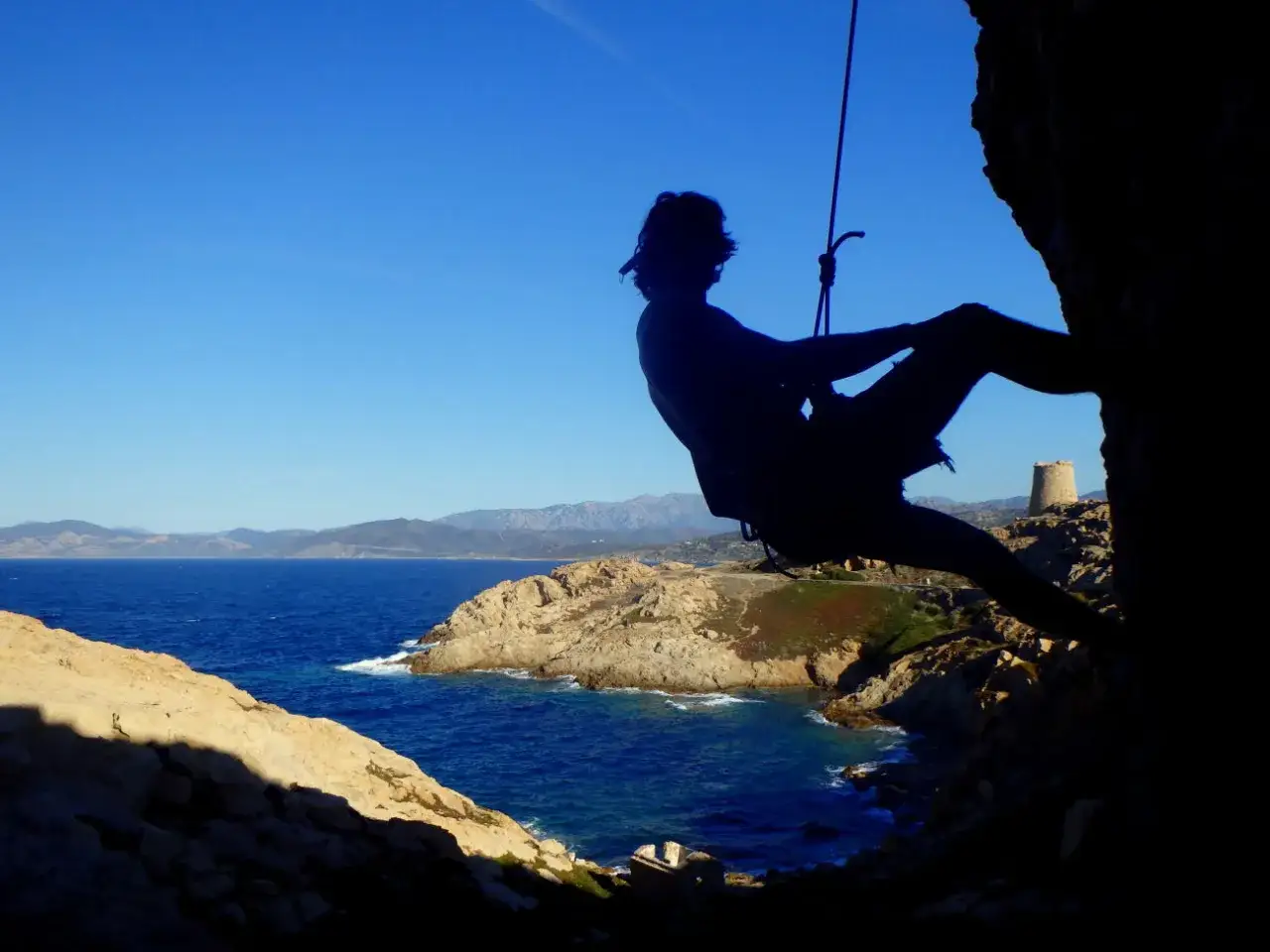 Silhouette eines Kletterers vor der K&uuml;ste Korsikas. Die Sonne scheint auf das Meer und die felsige Landschaft.