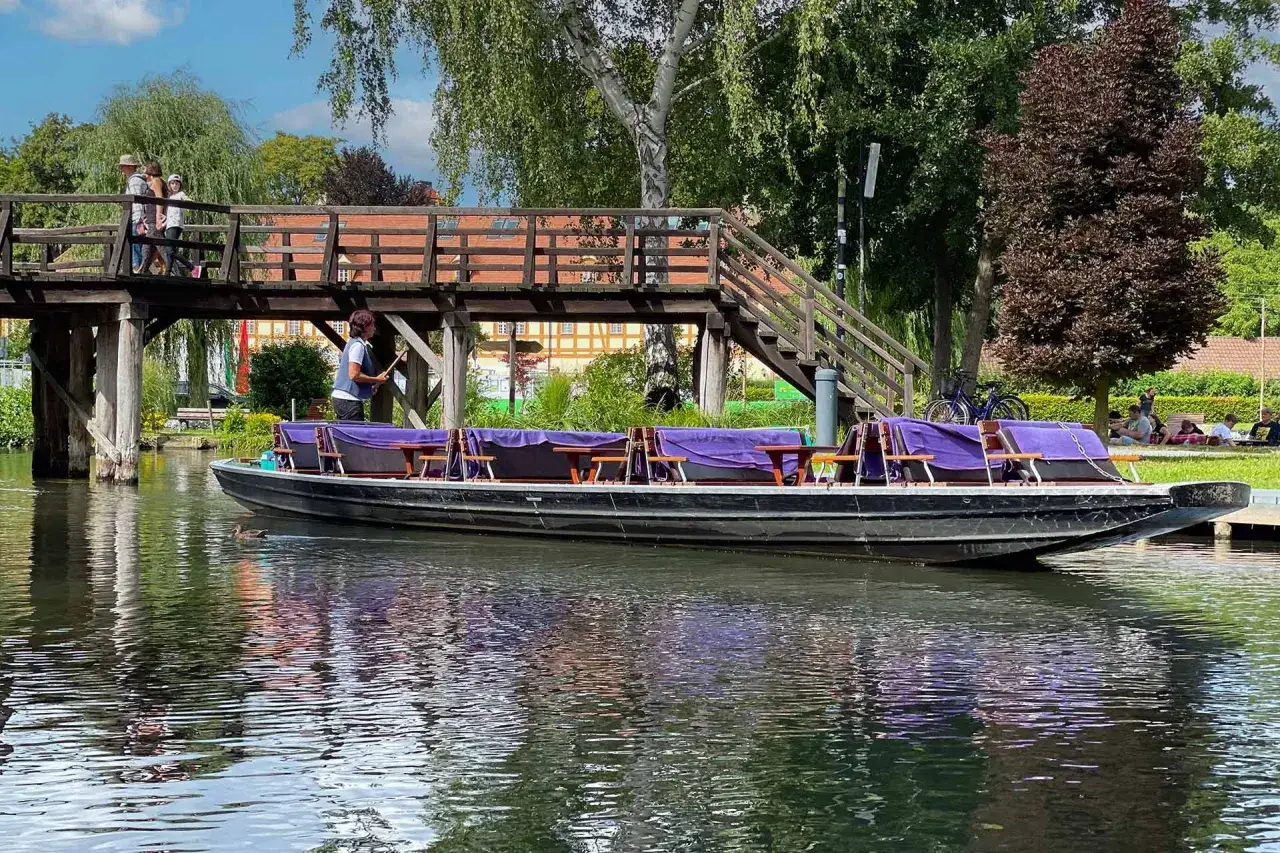 Kahnfahrt im Spreewald: Ein Ausflugsziel mit Charme. Ein Kahn mit violetten Bez&uuml;gen wartet auf G&auml;ste, w&auml;hrend Menschen &uuml;ber eine Holzbr&uuml;cke gehen.