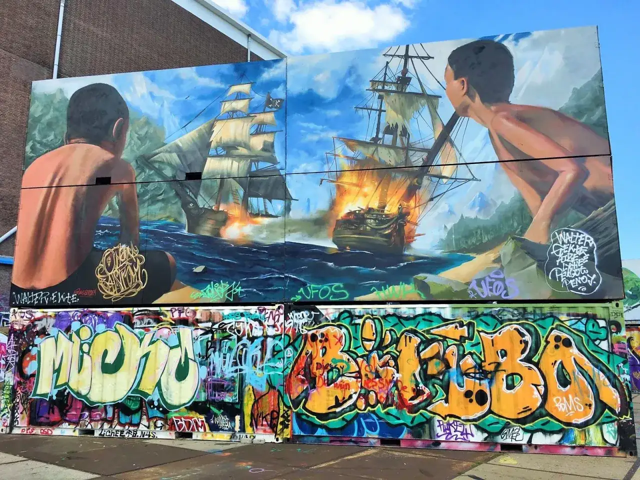 Graffiti-Kunst am NDSM in Amsterdam: Piratenschiffe in Flammen, beobachtet von zwei Jungen. Darunter bunte Schriftz&uuml;ge.
