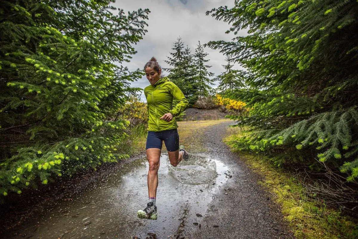 Frau im gr&uuml;nen Laufshirt und blauen Shorts macht slow jogging durch eine Pf&uuml;tze auf einem Waldweg.