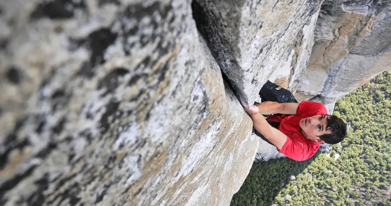 Ein Kletterer im roten Shirt hangelt sich an einer Felswand entlang, wie im free solo film. Tief unten erstreckt sich ein gr&uuml;ner Wald.