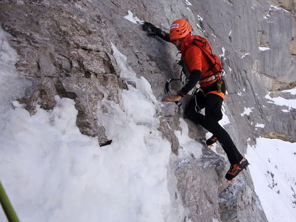 Ueli Steck klettert eine eisige Felswand hoch. Seine orangefarbene Kleidung und sein Helm heben sich vom grauen Fels ab.