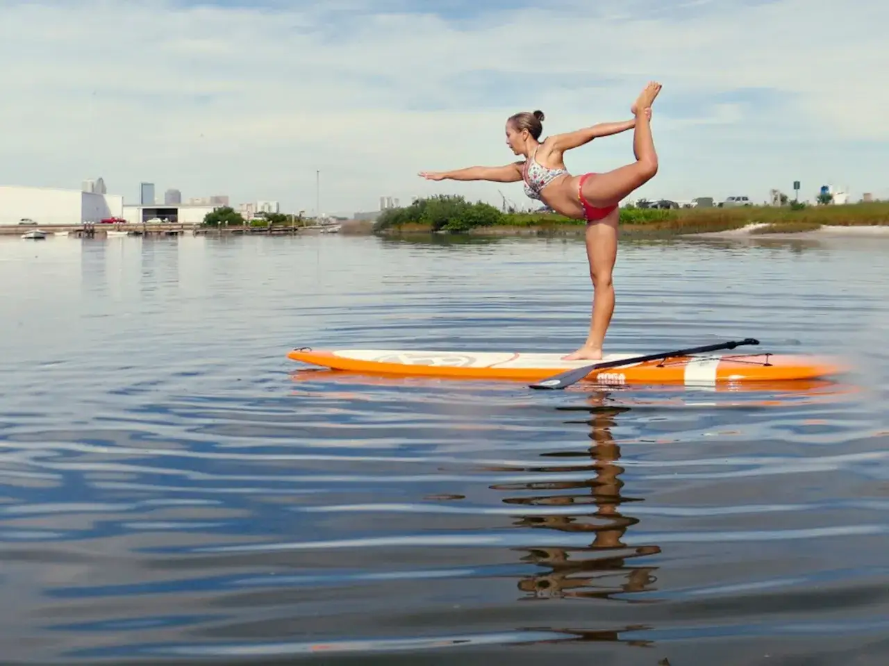 Frau in Bikini macht Krieger-Pose auf SUP-Board. Sup Yoga mit Blick auf Stadtkulisse.